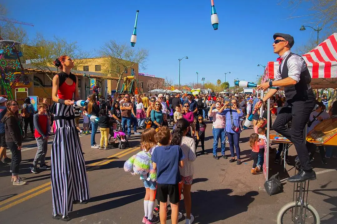 Tabitha on stilts and Dan on a unicycle juggling above children at an outdoor community event