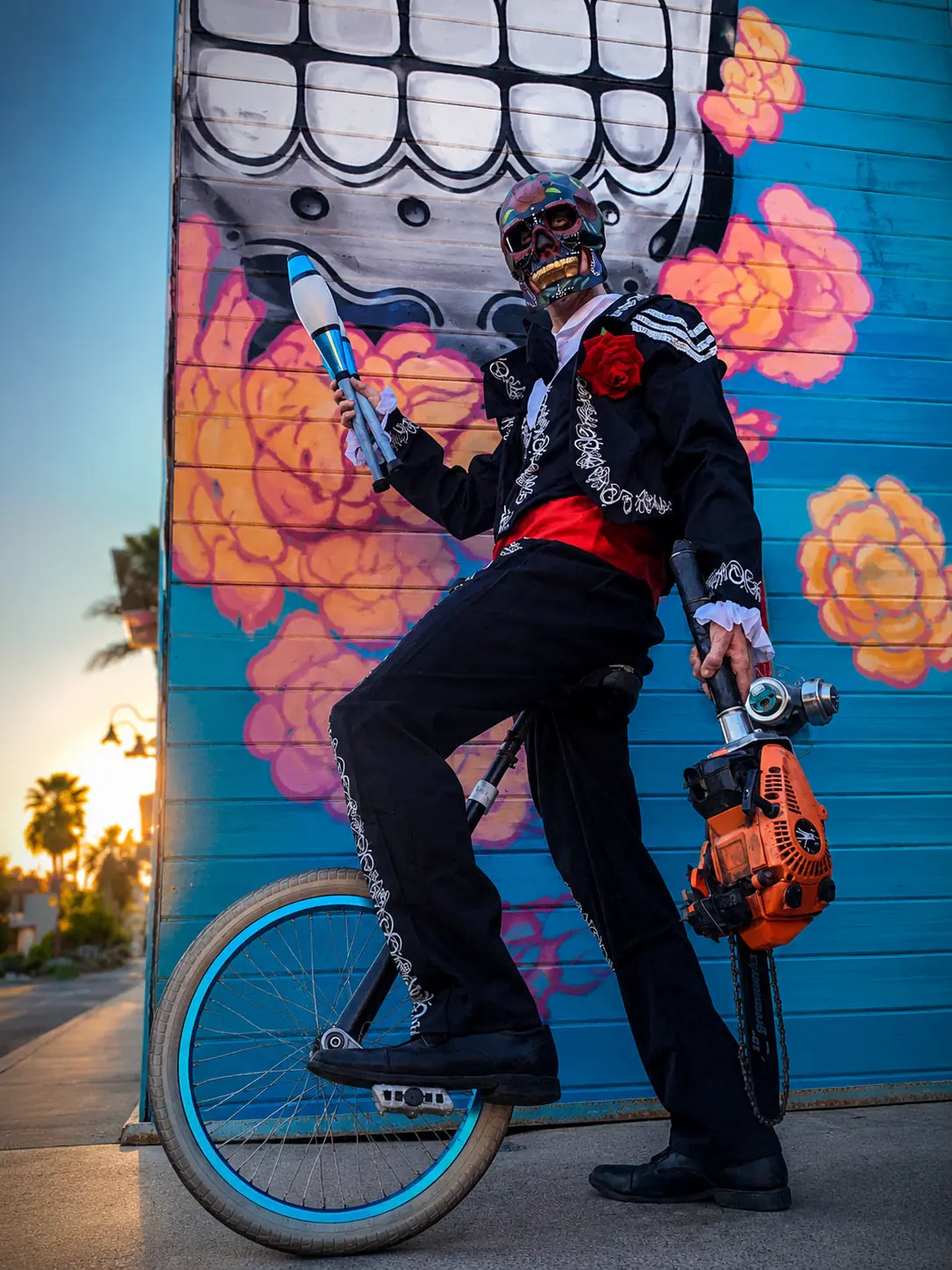 Dan in Day of the Dead costuming balancing on a unicycle with juggling clubs and a chainsaw