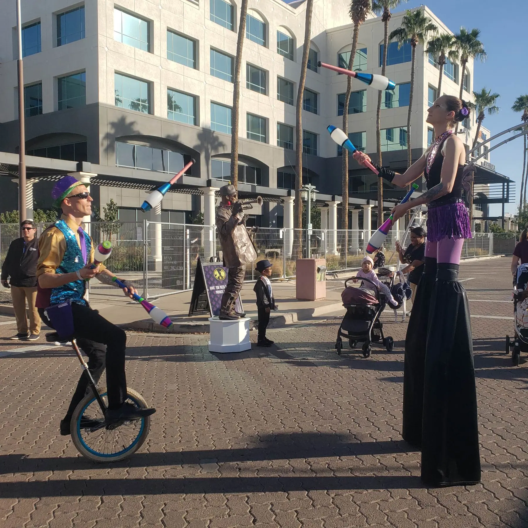 Dan riding a unicycle and Tabitha on stilts while they pass juggling clubs at a street festival
