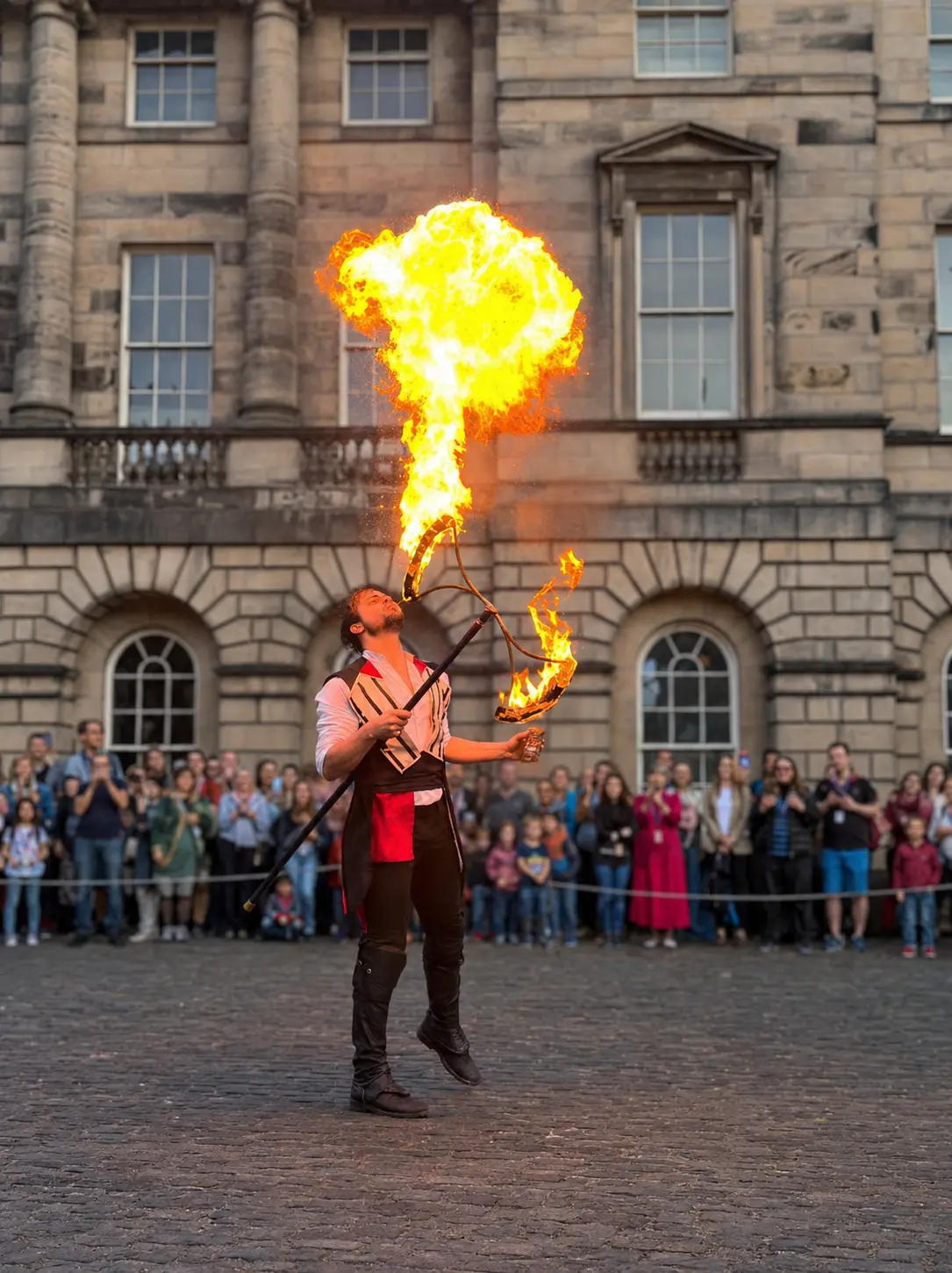 Andrew breathing a large flame while holding fire props at an outdoor festival