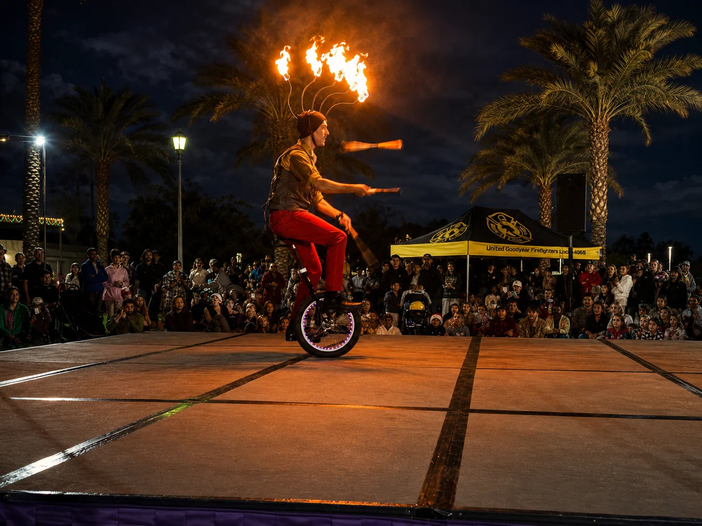 Dan balancing on a unicycle beneath a flaming headdress while juggling clubs on an outdoor stage