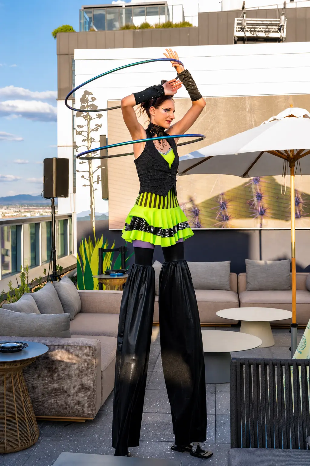 Tabitha hula hooping on stilts in a bright green circus costume at a daytime rooftop event