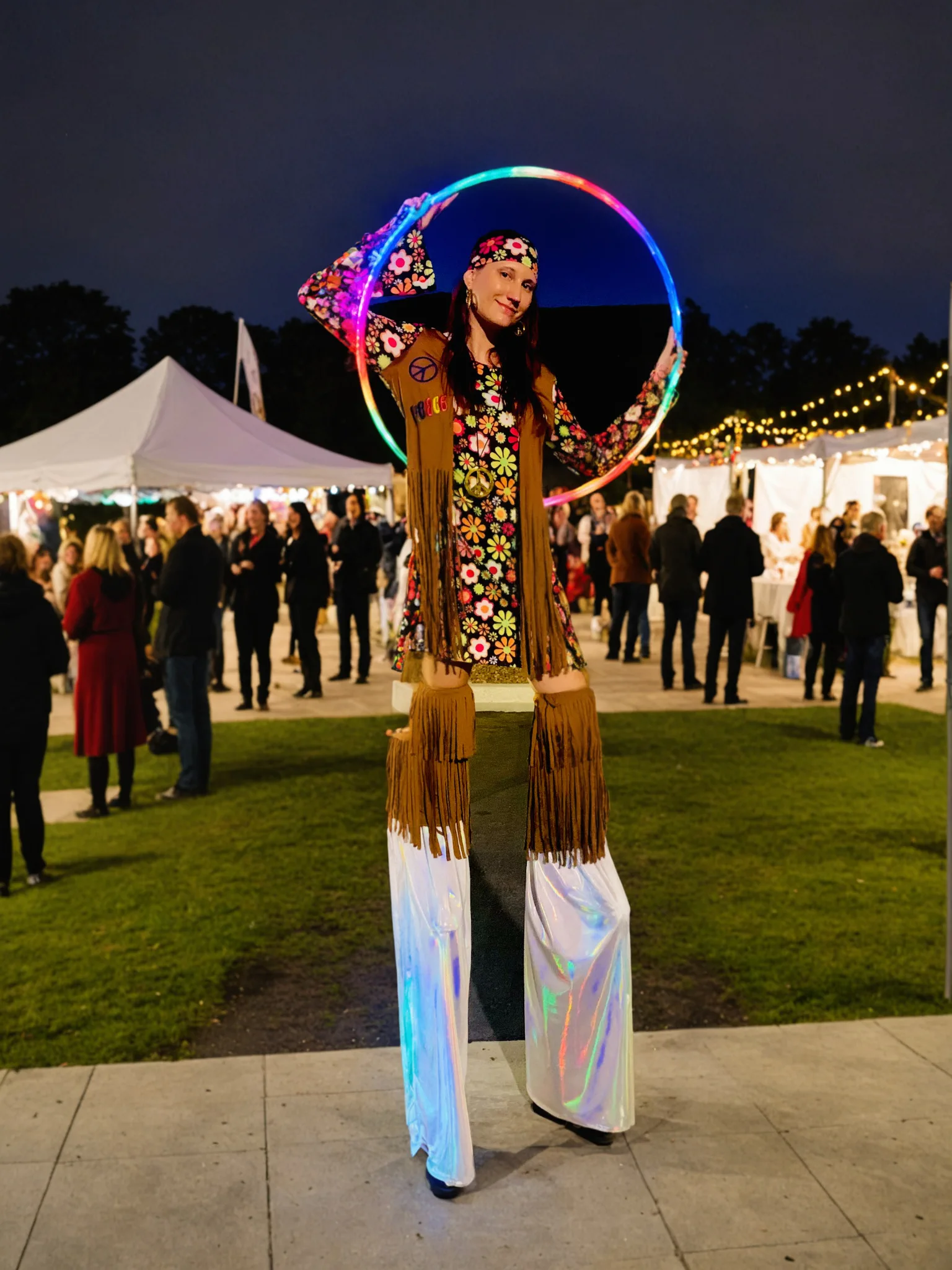 Tabitha on stilts in a 1960s hippie costume posing with an illuminated hoop