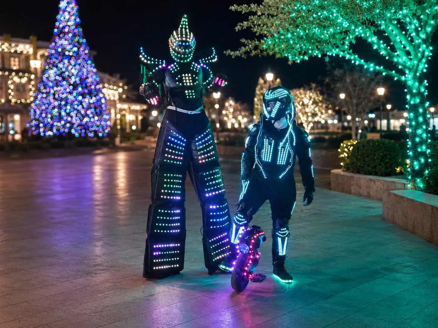 An LED stilt robot and illuminated jumping-stilt performer posing at a nighttime holiday event