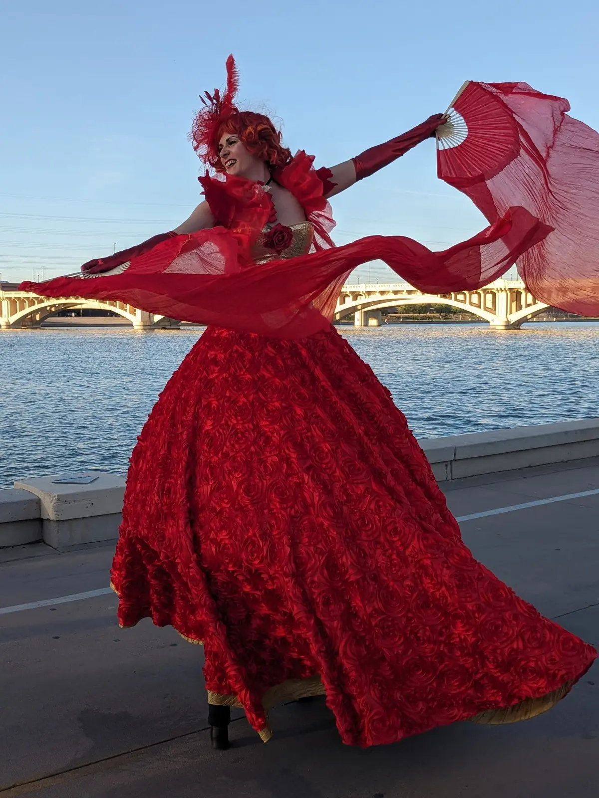A red-costumed stilt performer posing with flowing fans beside the water